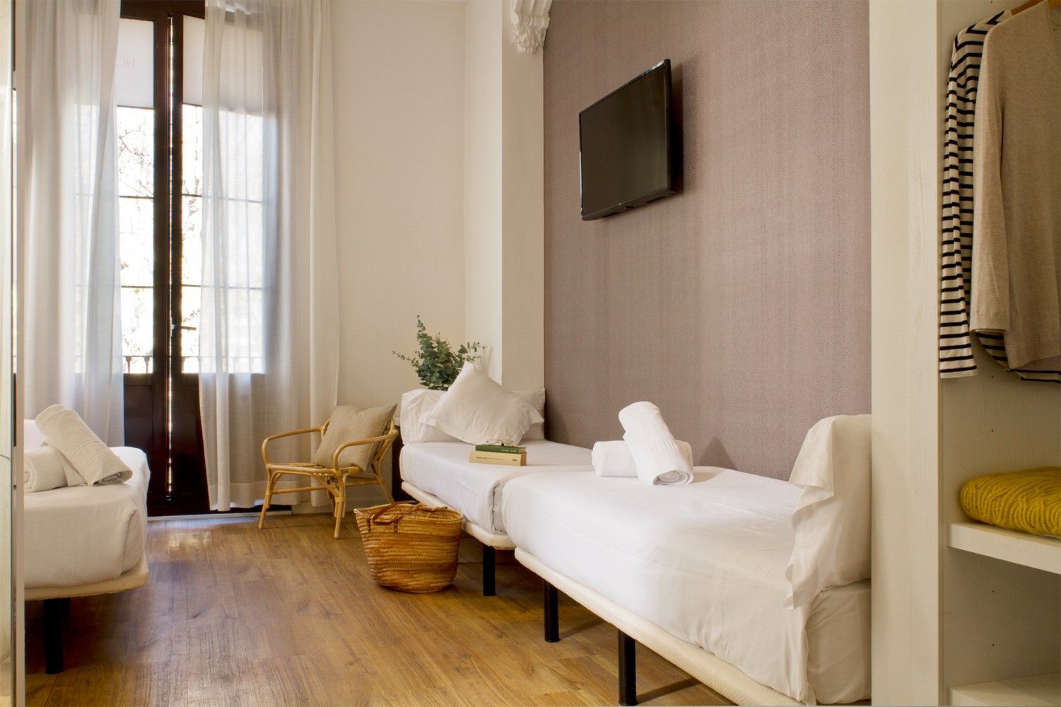 Una habitación de hotel con dos camas individuales blancas, una silla de ratán, una ventana con cortinas.