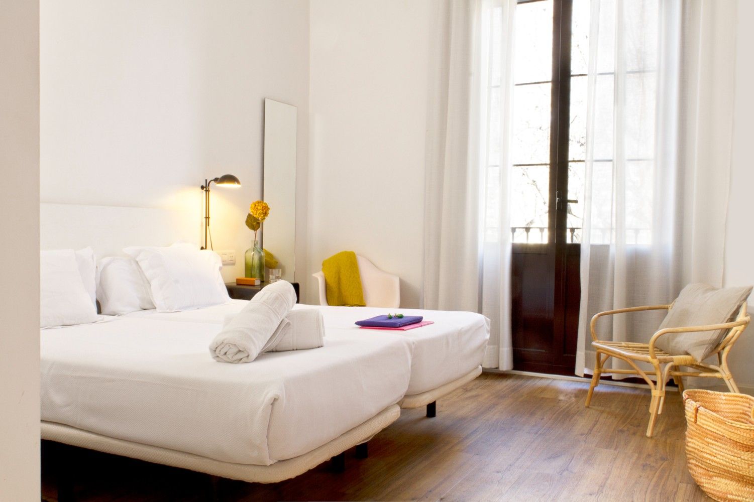 Un dormitorio de hotel minimalista con dos camas individuales blancas, una silla de ratán y cortinas transparentes.