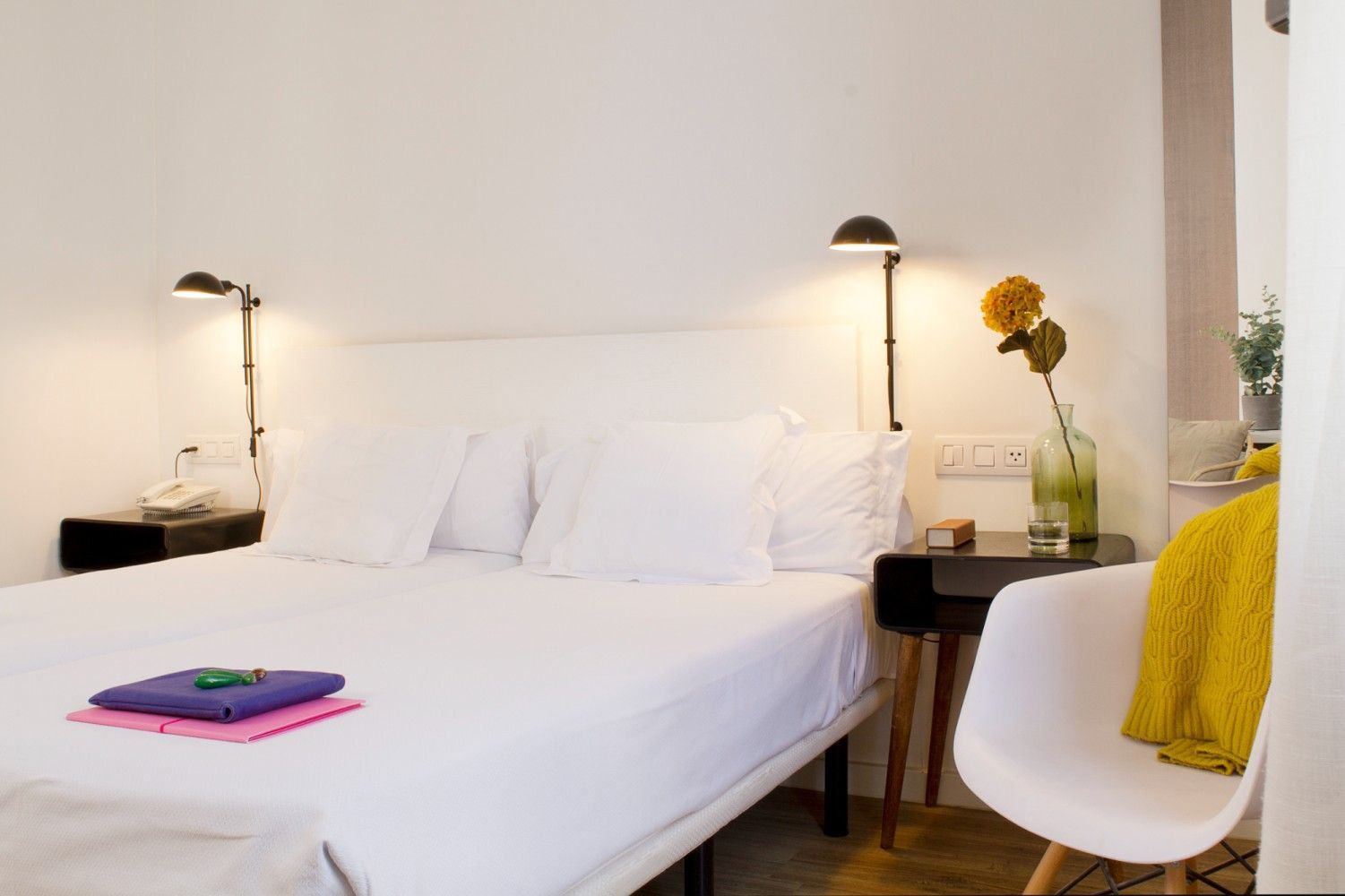 Una habitación de hotel con paredes blancas, una cama cuidadosamente hecha, mesas de noche negras con lámparas y una silla.