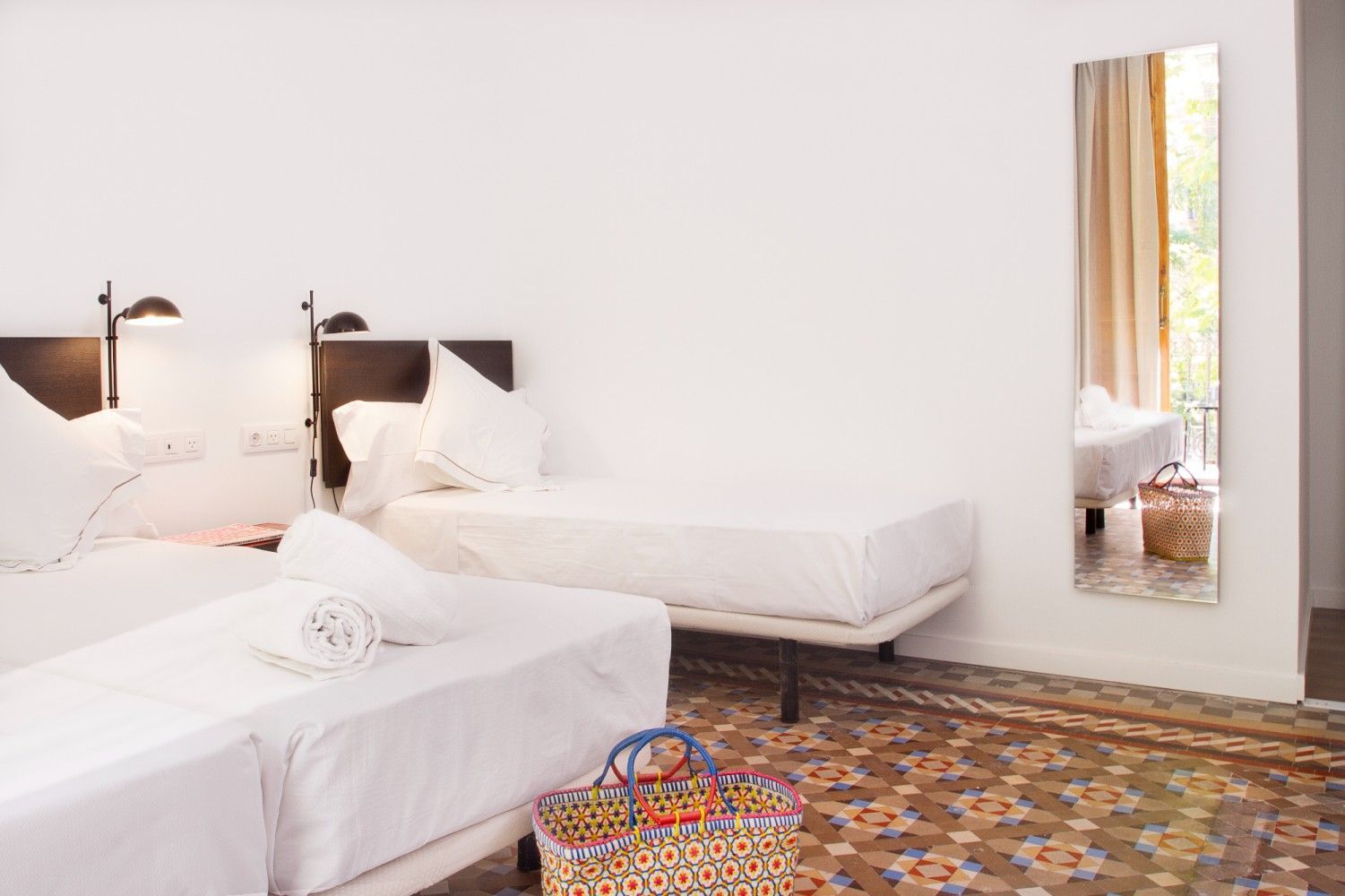 Dos camas individuales en una luminosa habitación de hotel con suelo de baldosas decorativas, un espejo de cuerpo entero.