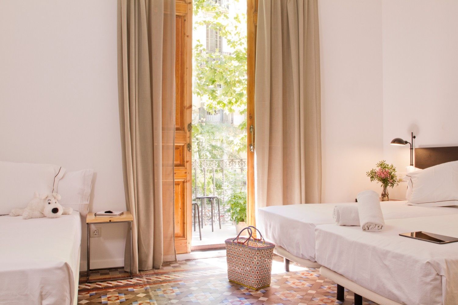 Un dormitorio luminoso con dos camas blancas, cortinas beige, una puerta abierta a un balcón.