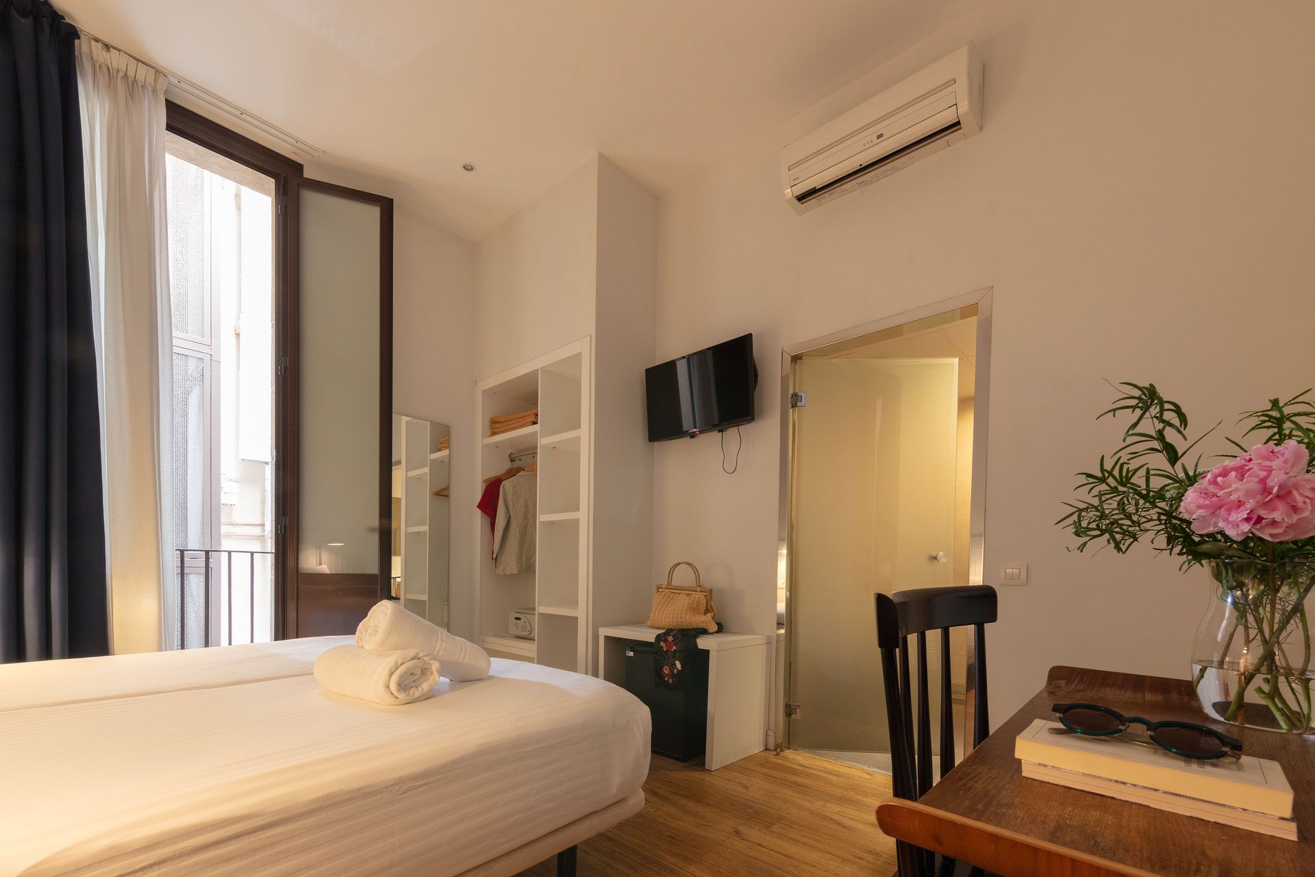 Habitación luminosa con cama, puerta que da al balcón, televisor de pared, aire acondicionado y una mesita con flores.