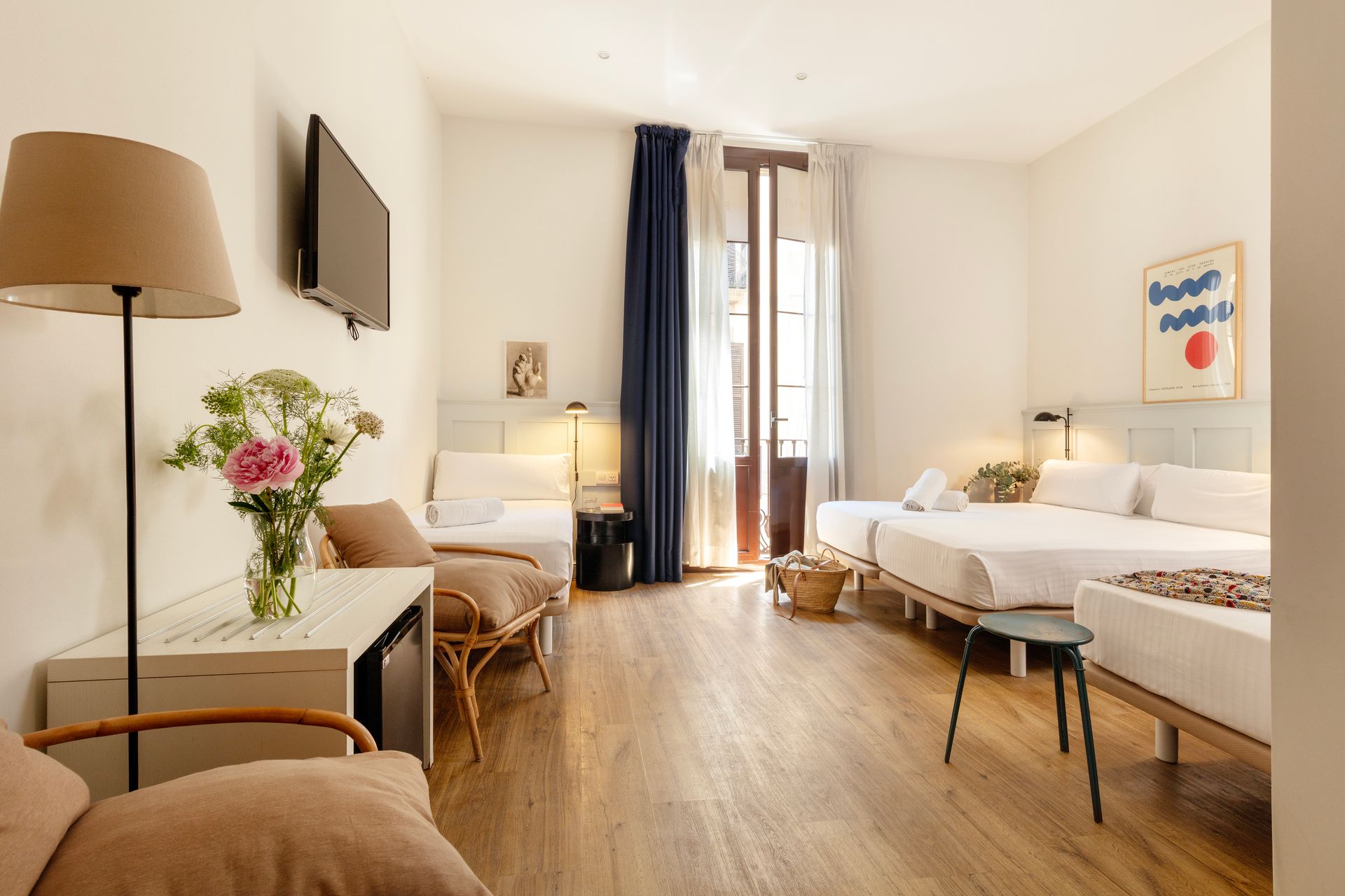 Habitación de hotel luminosa con dos camas individuales, suelos de madera, ventanas altas y una mesa con flores.