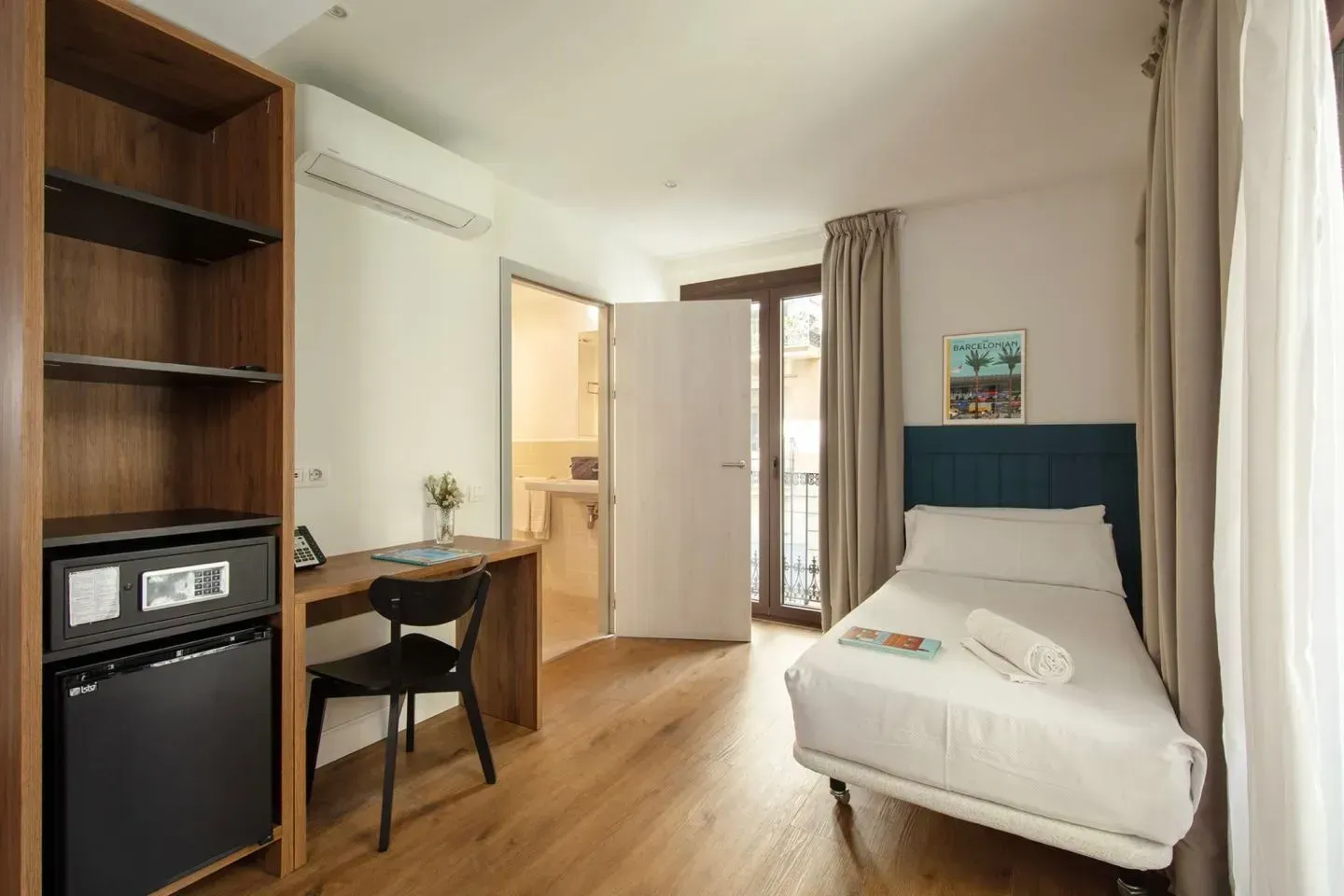 Una habitación de hotel con una cama individual, un escritorio con una silla negra, un espacio de almacenamiento abierto.