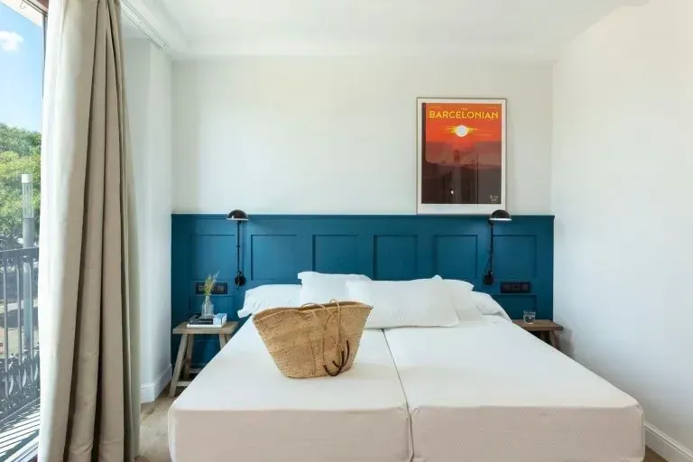 Un dormitorio minimalista con una cabecera con paneles de color verde azulado, ropa de cama blanca, un bolso de paja.