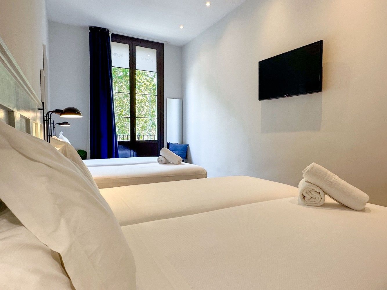 Una luminosa habitación de hotel con dos camas blancas, paredes blancas, un televisor montado en la pared.