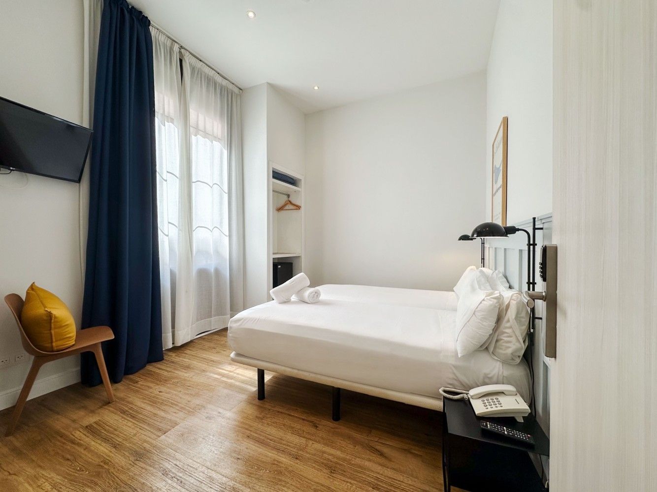 Una habitación de hotel con una cama blanca cuidadosamente hecha, pisos de madera, cortinas azul marino, una silla amarilla.