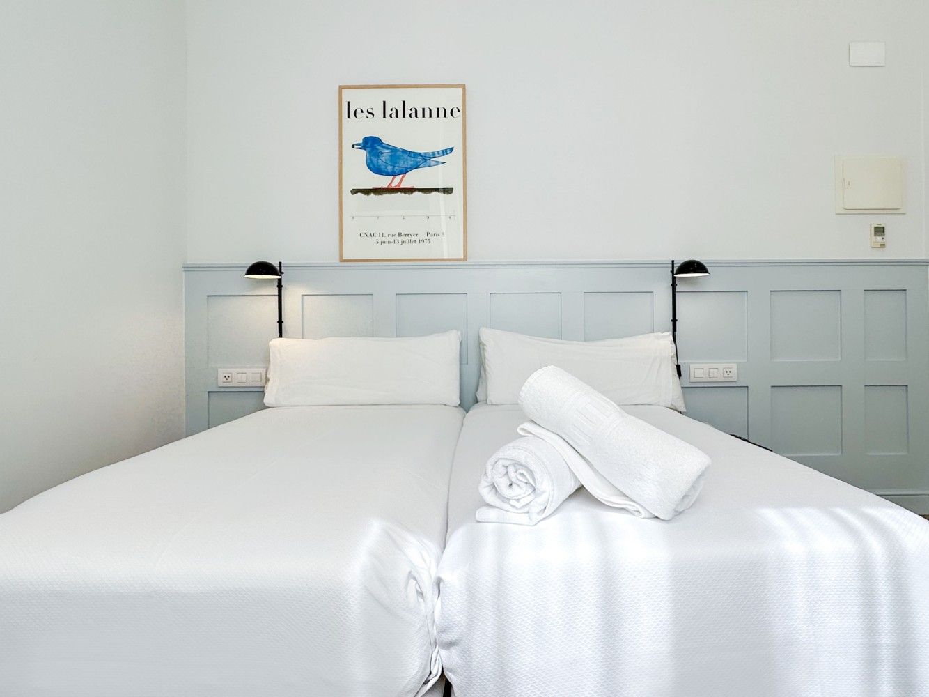 Dos camas individuales con ropa de cama blanca impecable y toallas apiladas, frente a una pared con paneles de madera .