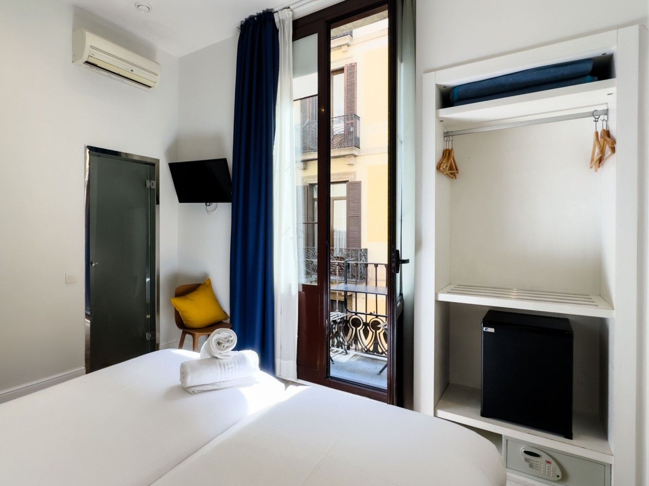 Una habitación de hotel limpia con una cama, sábanas blancas, una almohada amarilla, una nevera pequeña.