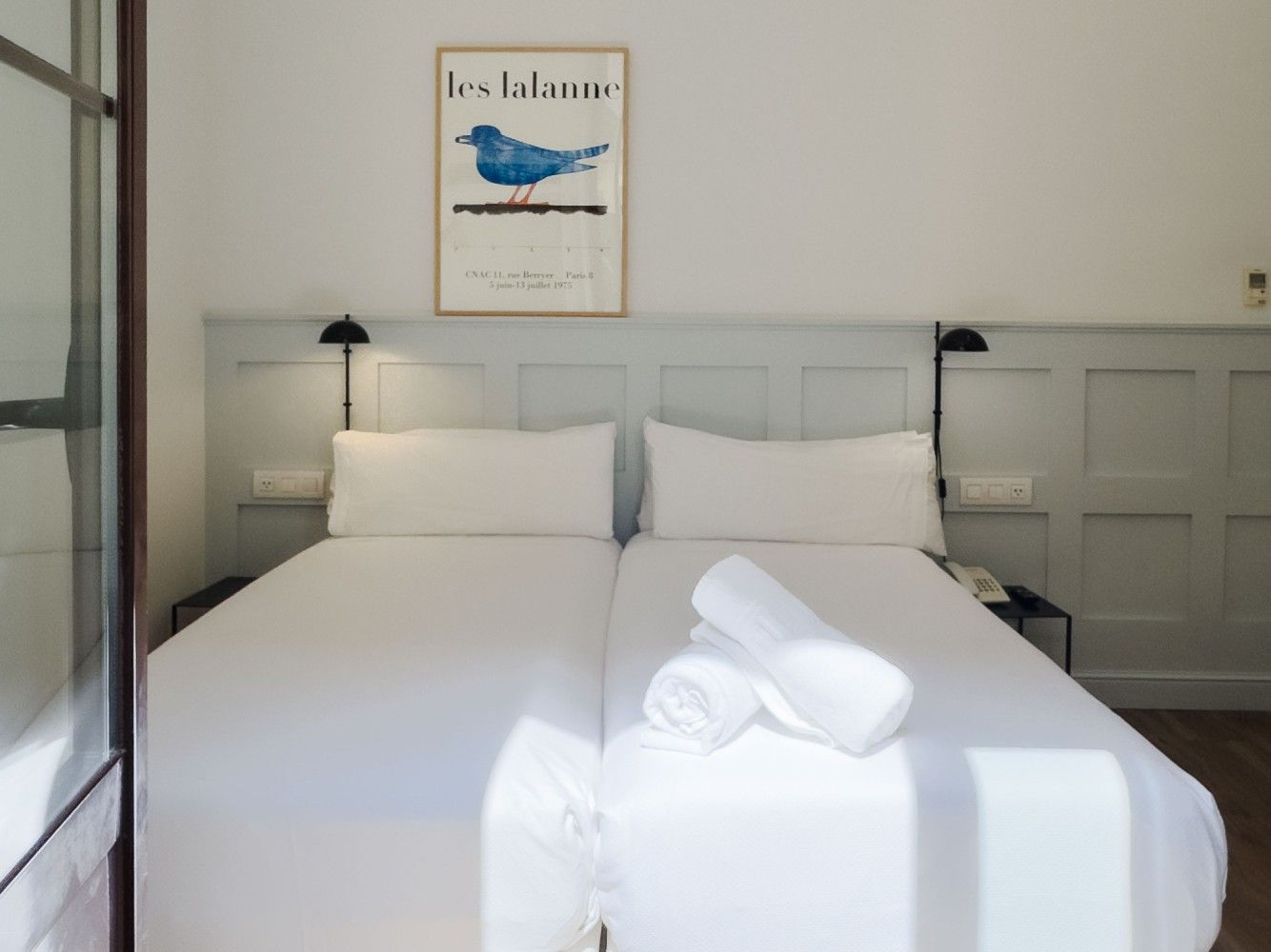 Una habitación de hotel con dos camas individuales blancas, revestimiento de madera gris claro y un cuadro con un pájaro.