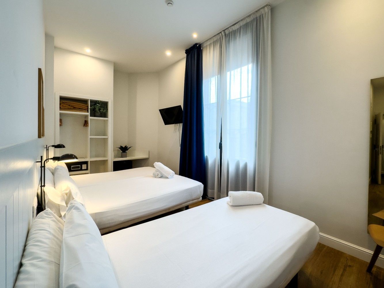 Una moderna habitación de hotel con dos camas blancas, paredes blancas y ventanas de piso a techo con cortinas azul oscuro.