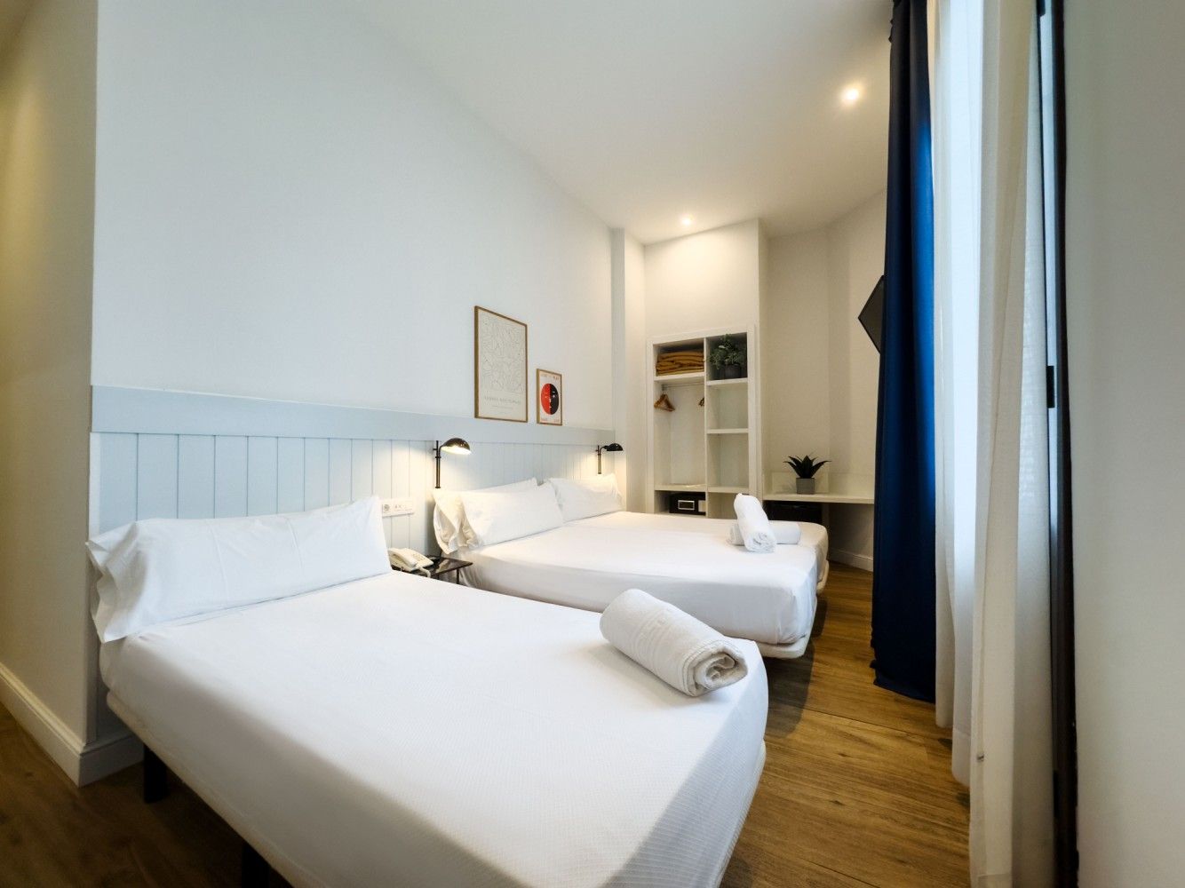 Una habitación de hotel con dos camas blancas, suelos de madera clara, paredes blancas y una pequeña estantería.