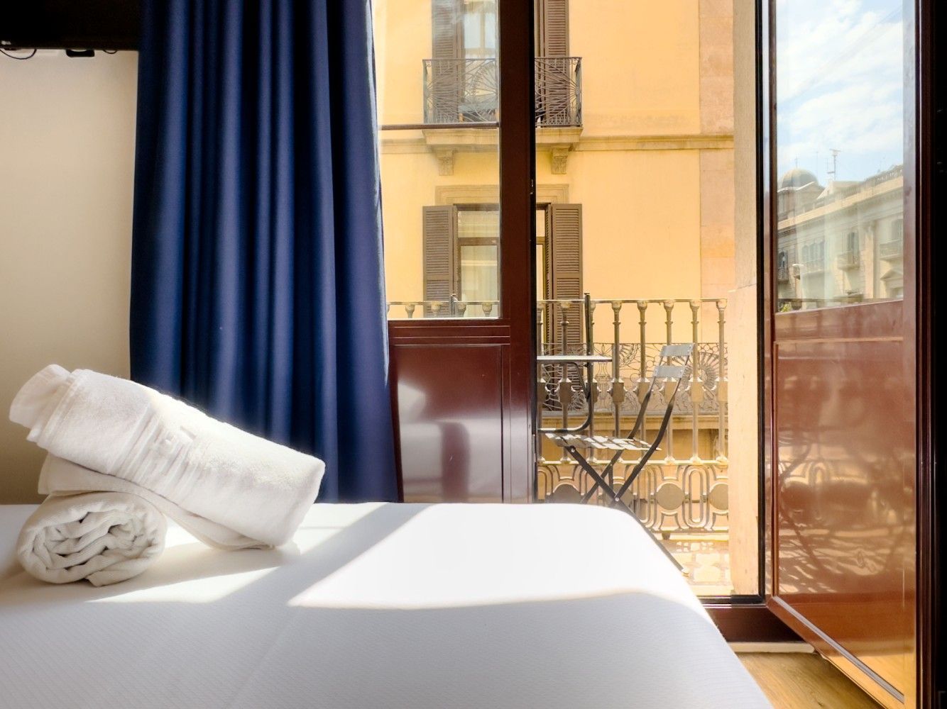 Toallas blancas dobladas están sobre una cama junto a una ventana que da a un balcón iluminado por el sol.