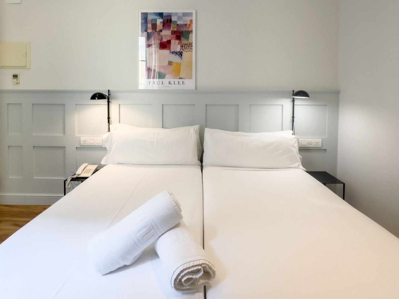 Dos camas individuales juntas con ropa de cama blanca, toallas enrolladas, lámparas de lectura y arte de pared .