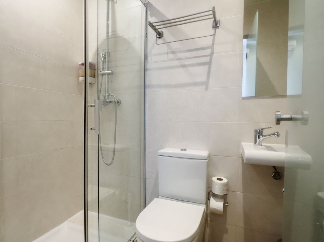 Pequeño baño con inodoro blanco, cabina de ducha de cristal, lavabo montado en la pared.