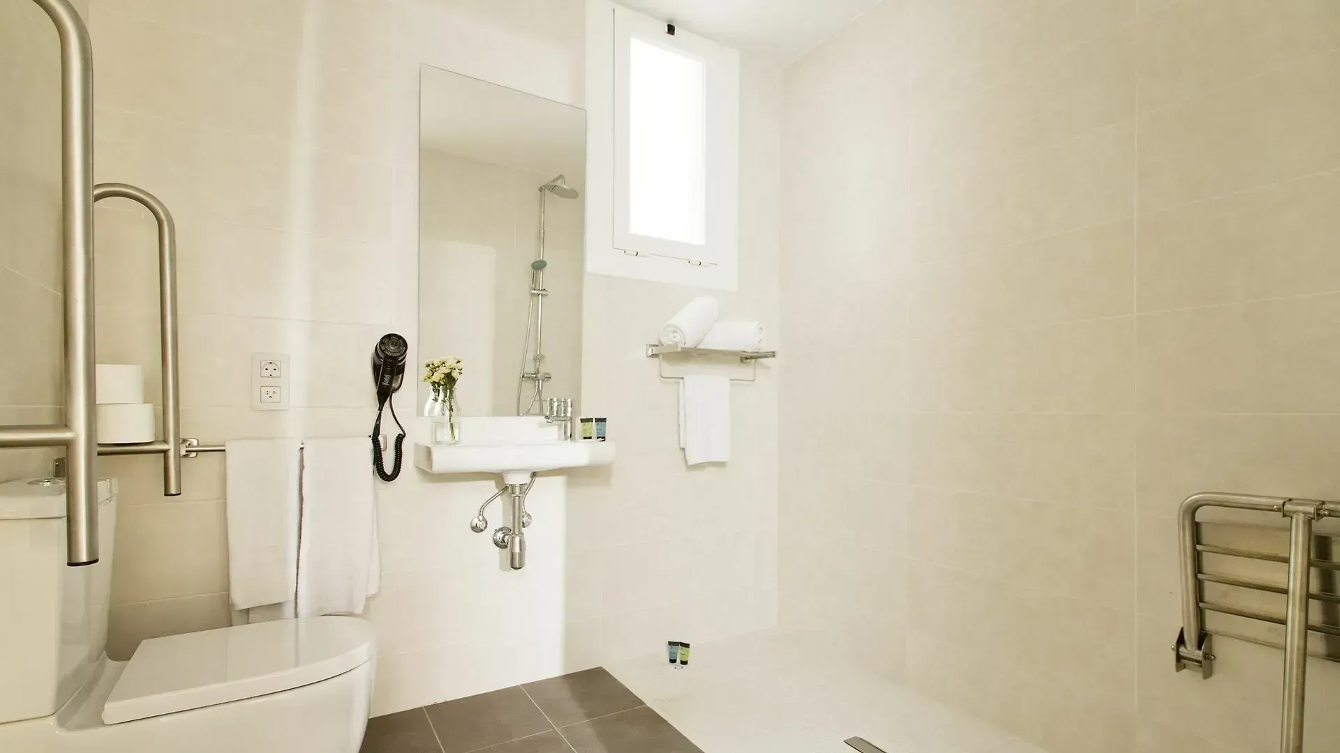 Un baño de hotel accesible con inodoro blanco, lavabo con espejo, ducha y barras de apoyo.