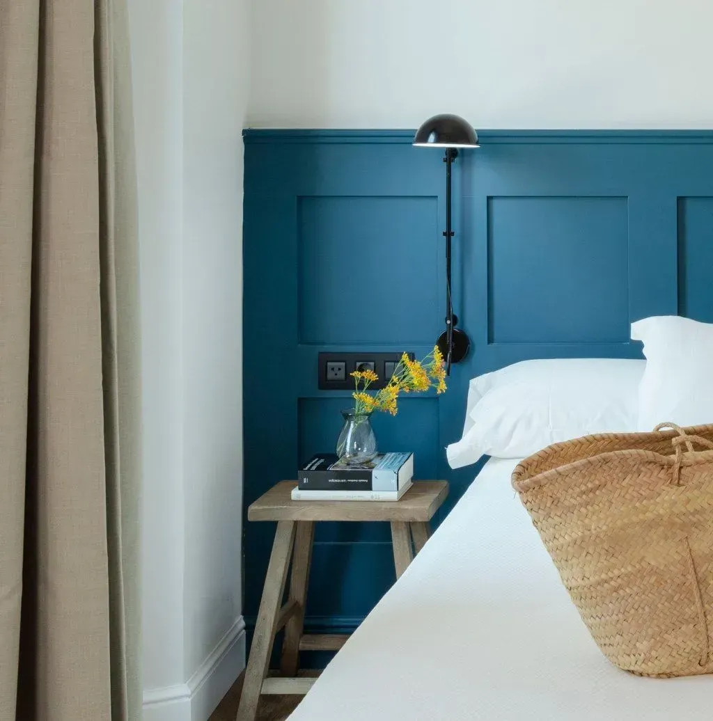 Un rincón de dormitorio con un cabecero con paneles azules, un pequeño taburete de madera como mesita de noche.