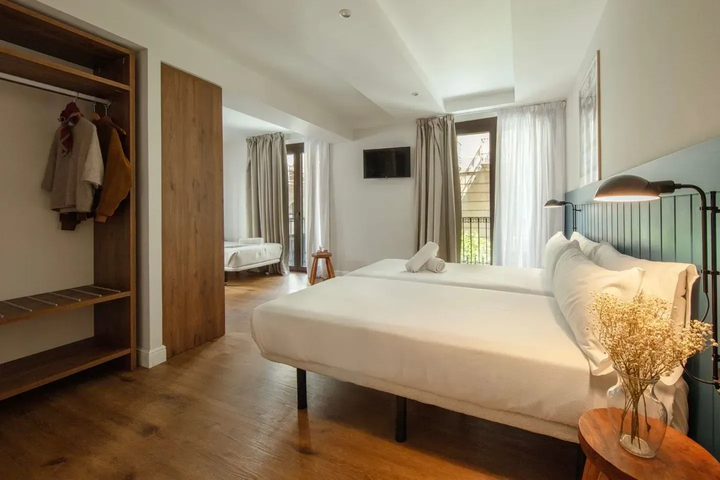 Una habitación de hotel con dos camas individuales, un armario abierto de madera y una pequeña mesa auxilia.