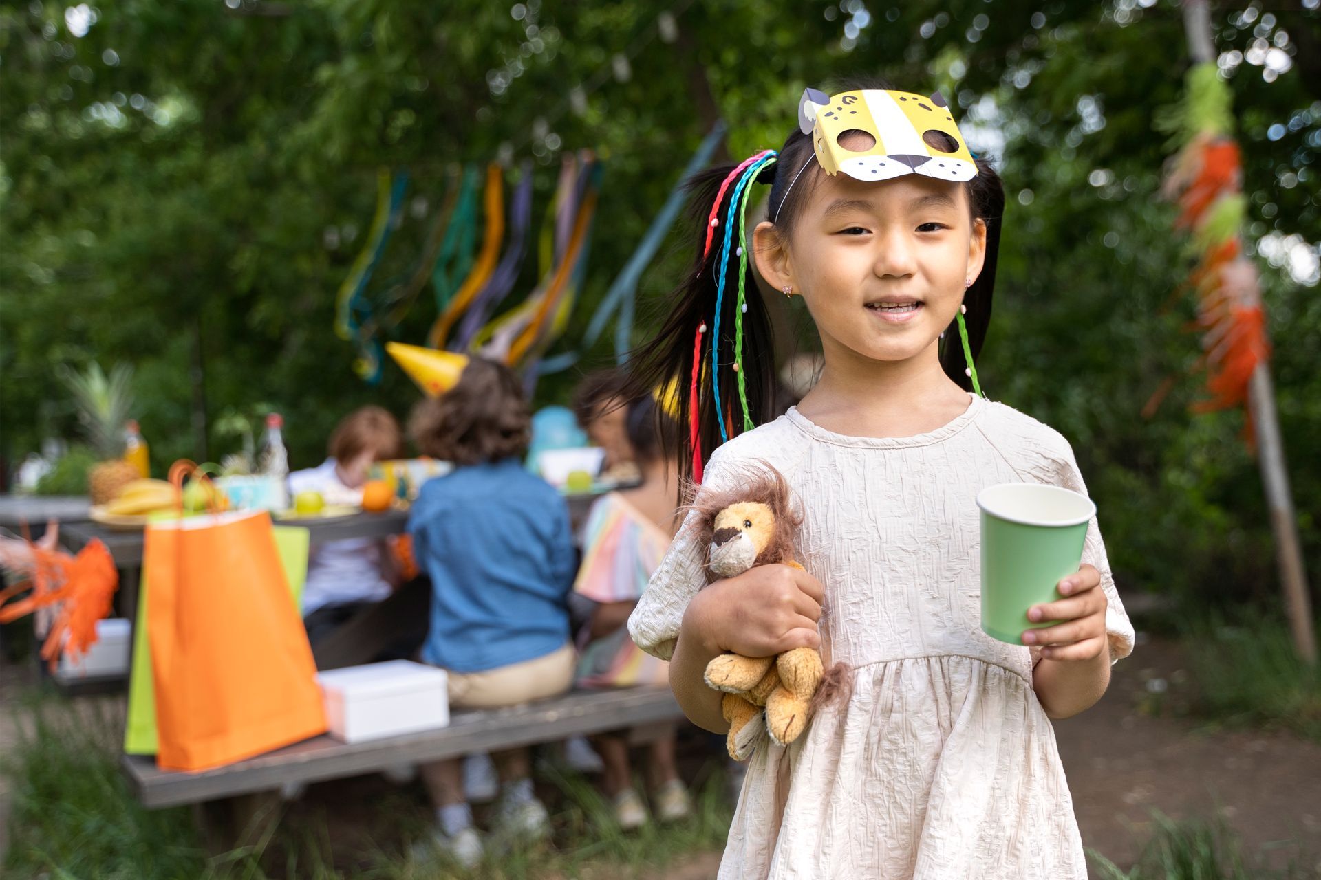 A little girl wearing a mask is holding a teddy bear and a cup at a birthday party.