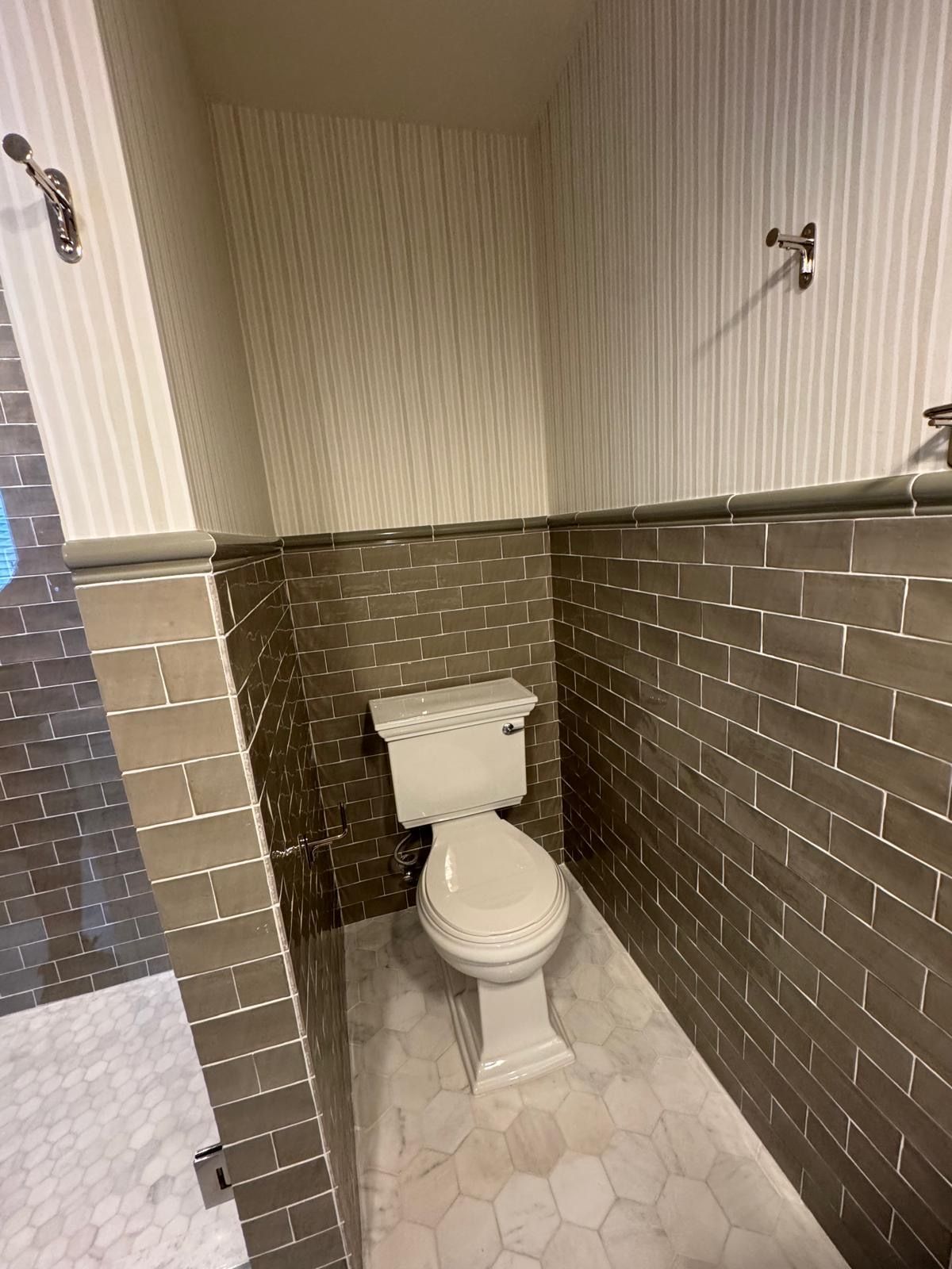 Narrow bathroom with a white toilet and hexagonal floor tiles. Gray brick-like wall tiles. Striped wallpaper.