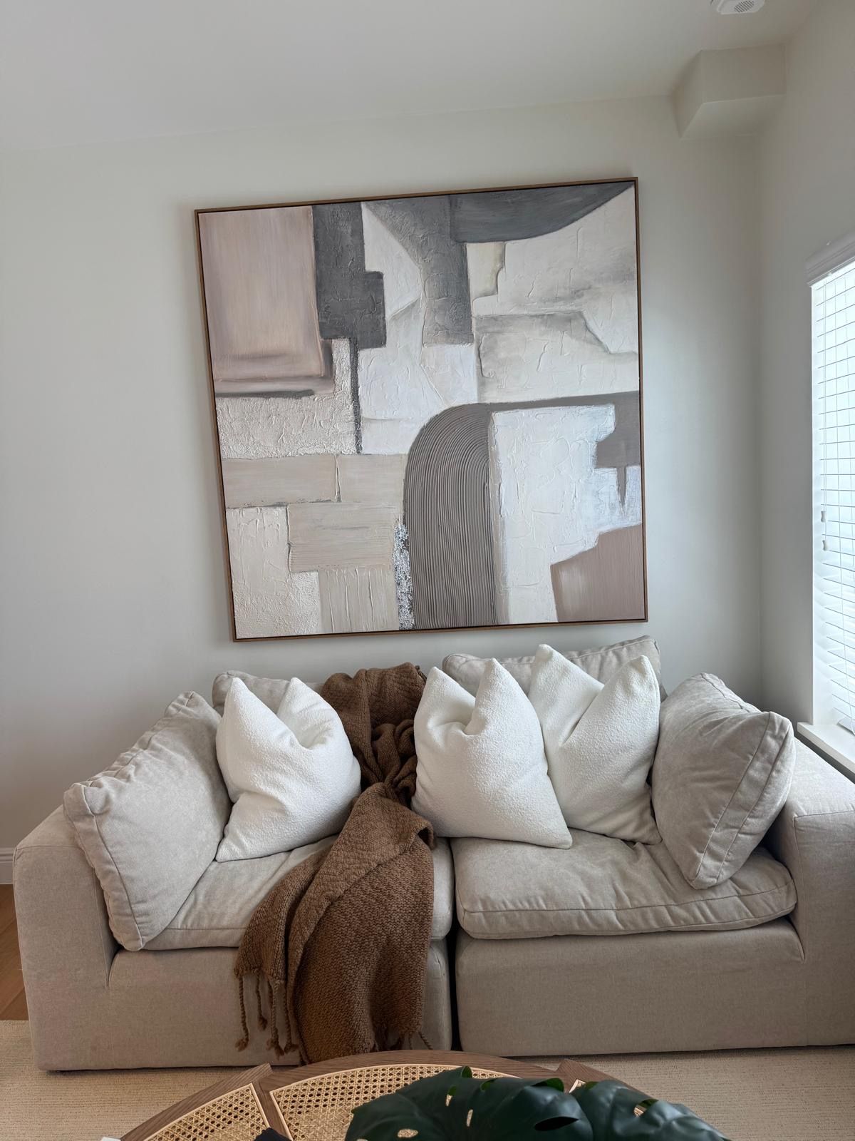 Beige sofa with white pillows and brown throw under abstract art in a neutral-toned living room.
