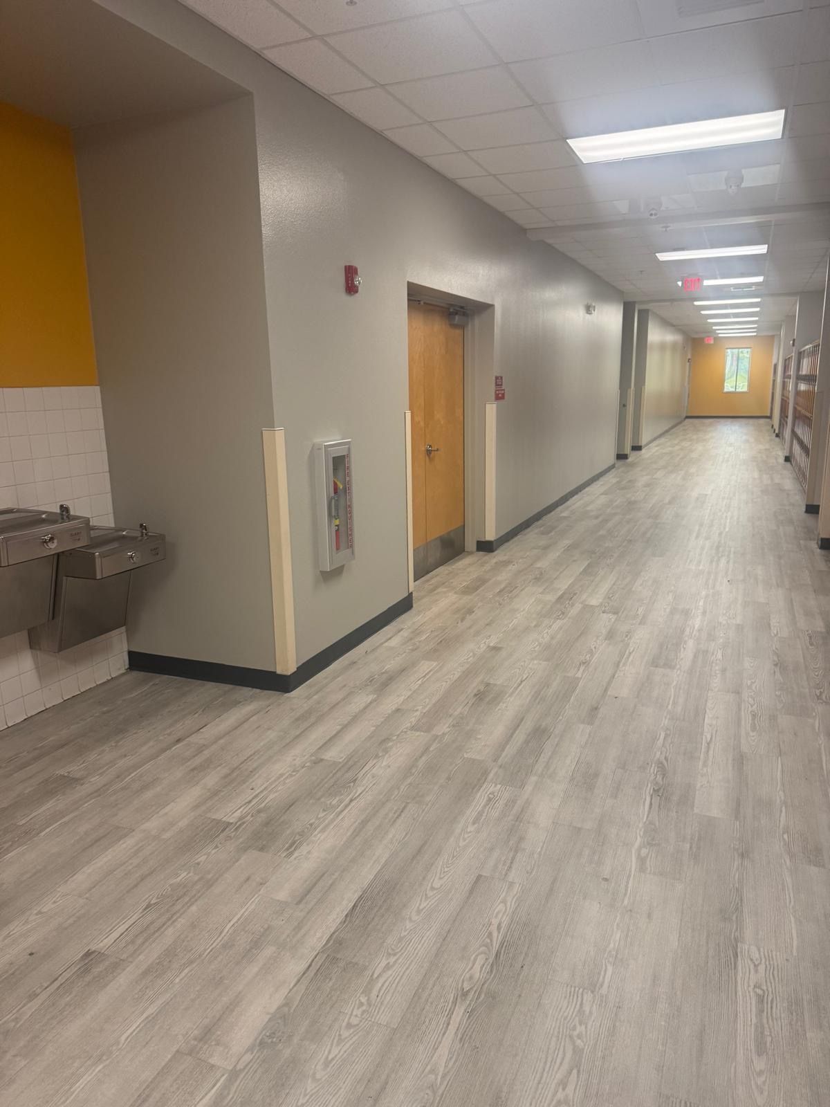 Hallway with light gray walls, wood-look flooring, yellow accent wall, water fountain, door, and fire extinguisher.