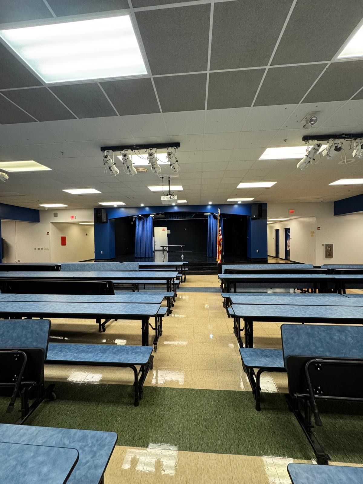 Empty school cafeteria with rows of blue tables facing a small stage.