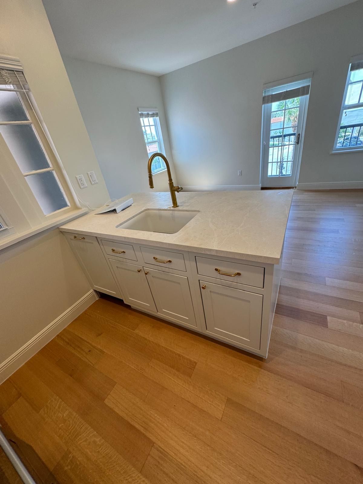 Kitchen island with sink, white cabinets, gold faucet, light wood floor. Natural light fills room.
