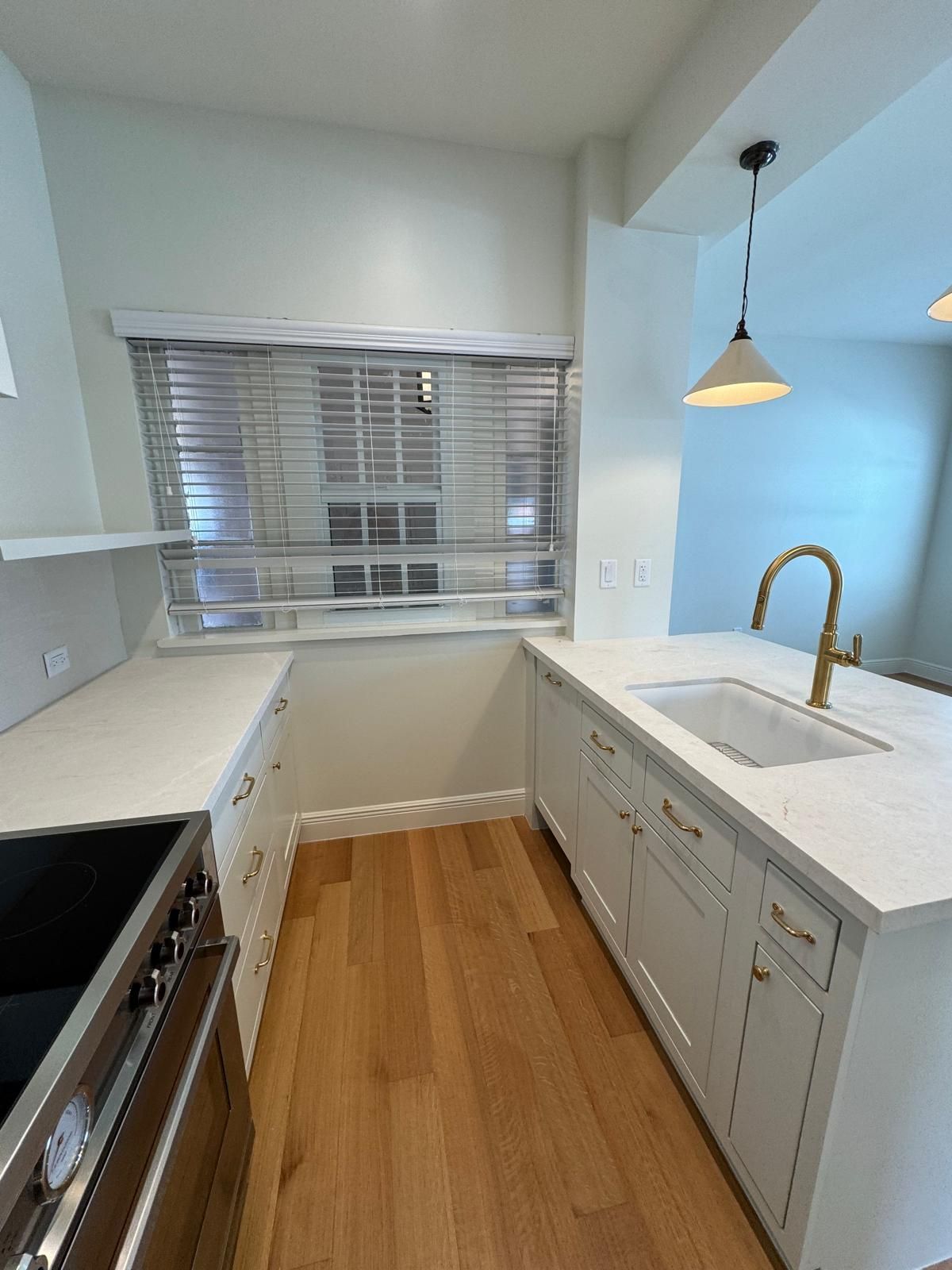 Kitchen with white cabinets, island with sink, hardwood floors, window with blinds, and gold faucet.