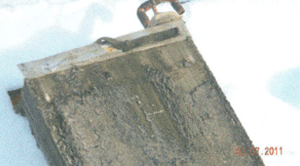 Close-up view of a rectangular, weathered, gray metal object with a handle attached at the top.