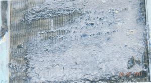 Close-up of a rectangular, metallic component heavily coated in thick, grey, dried residue.