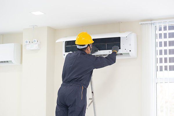 A worker in a yellow helmet cleans an air conditioning unit indoors, standing on a ladder. A worker in a yellow helmet cleans an air conditioning unit indoors, standing on a ladder.
