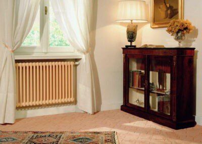 Radiator by a window with white curtains, next to a dark wood cabinet, flowers, and a lamp.