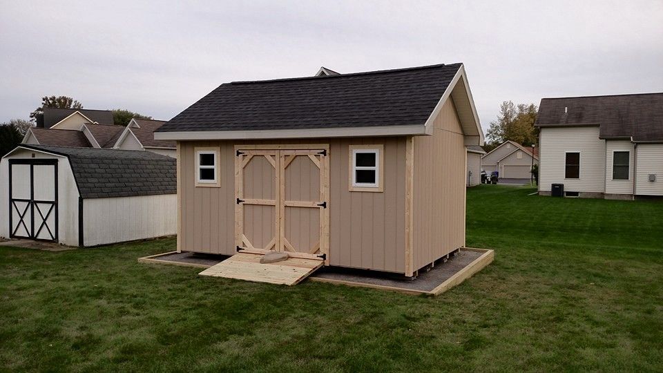 Tan shed with black roof, wooden double doors, and two small windows on a grassy backyard.