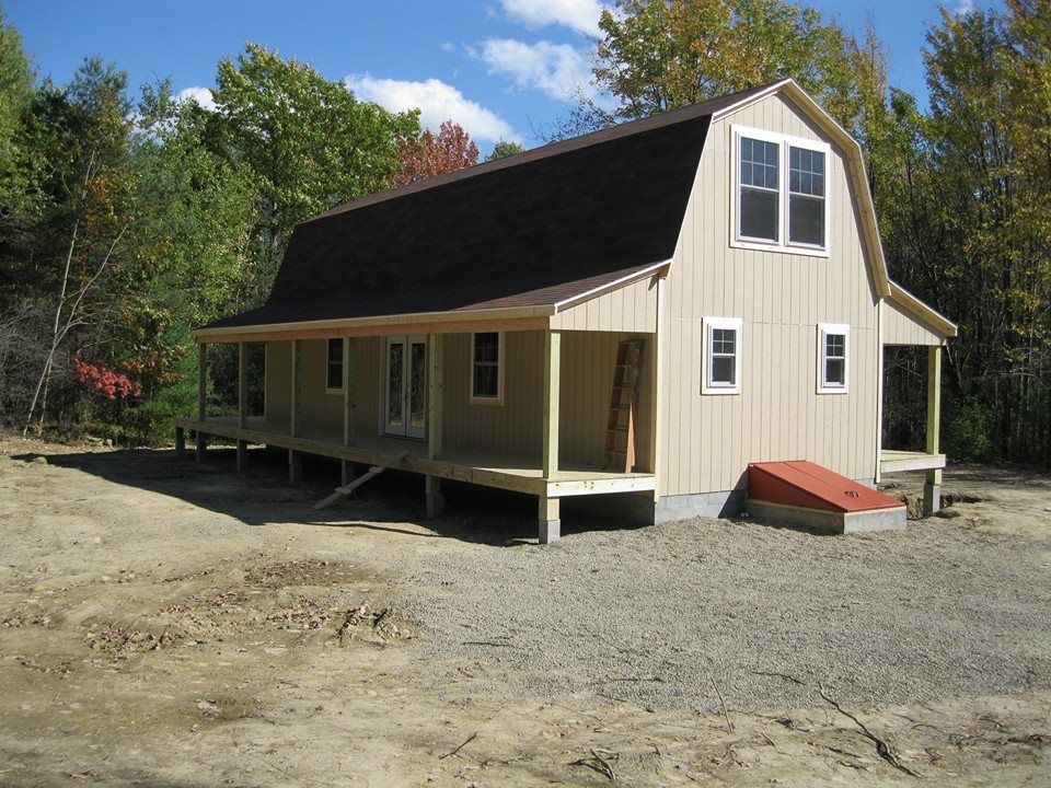 Beige Dutch-style house with brown roof and wraparound porch, set in a gravel lot surrounded by trees.