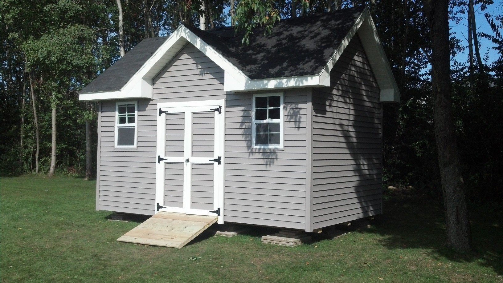 Gray shed with ramp in a grassy area, trees in the background. White trim and black roof.