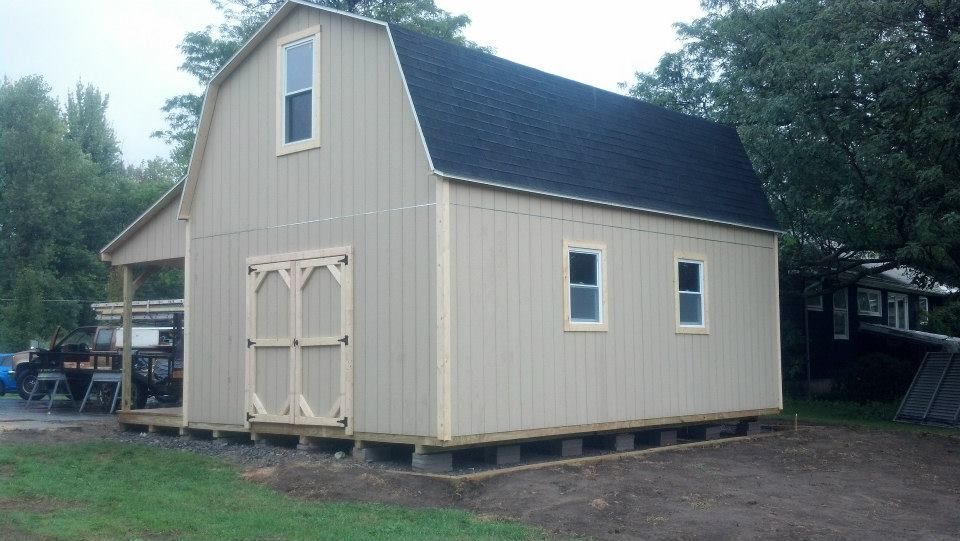 Tan barn with black roof, light wood trim, and a porch, on concrete blocks.