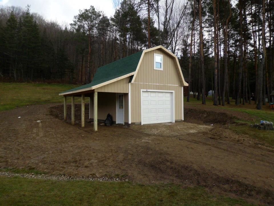 Tan barn with green roof, carport, and garage door on a dirt lot, surrounded by trees.