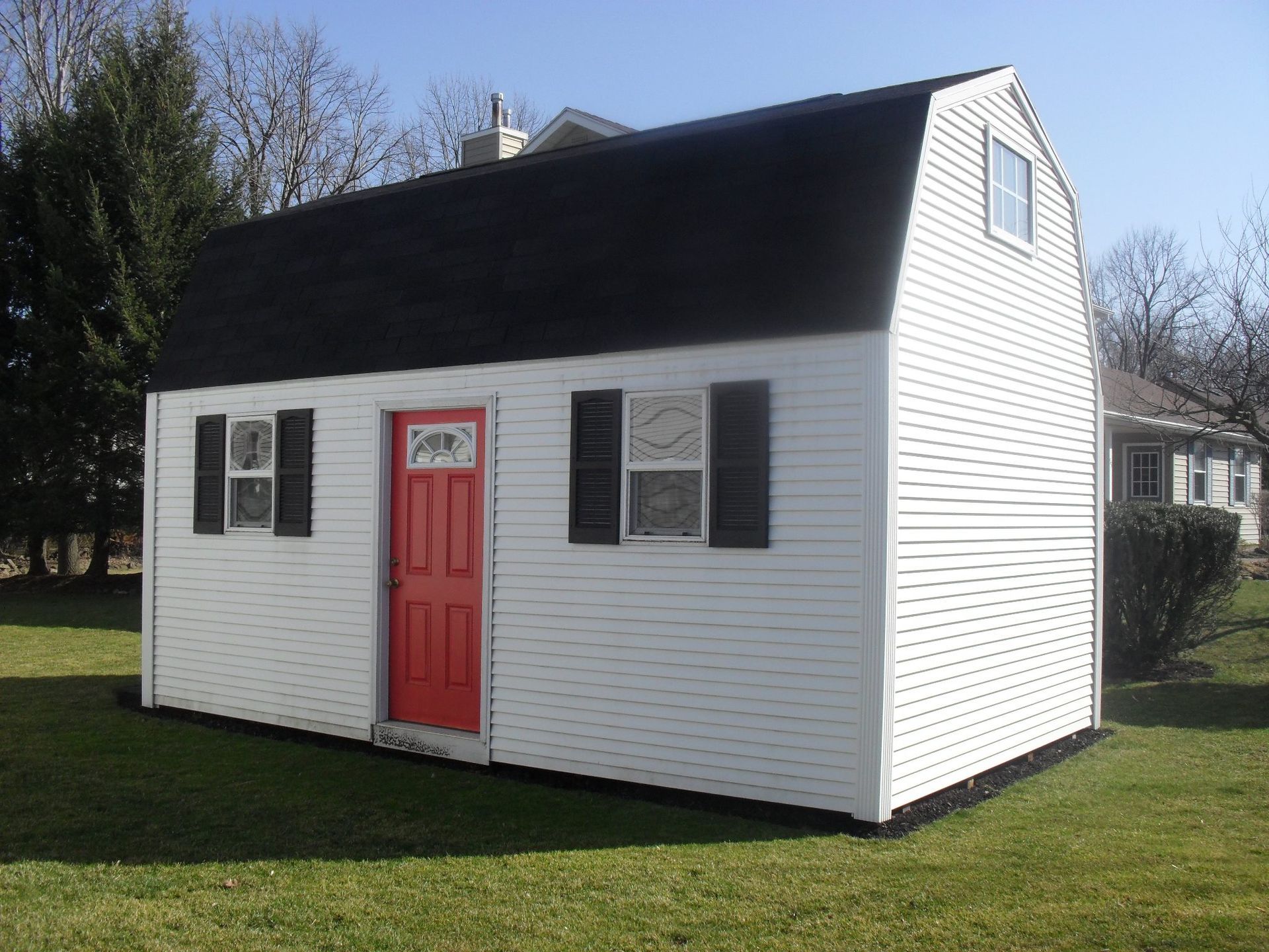 White shed with black roof, red door, and black shutters on windows, on a green lawn.