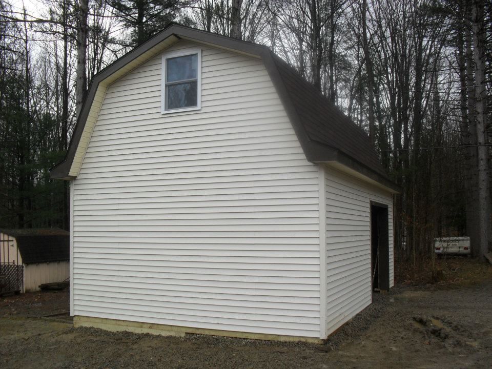 White barn-style shed with a dark roof and small window against a wooded backdrop.