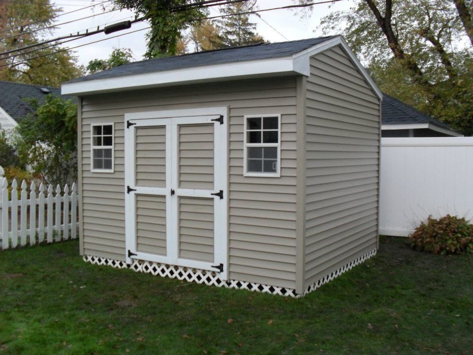 Tan storage shed with white trim, double doors, and windows, on a green lawn.