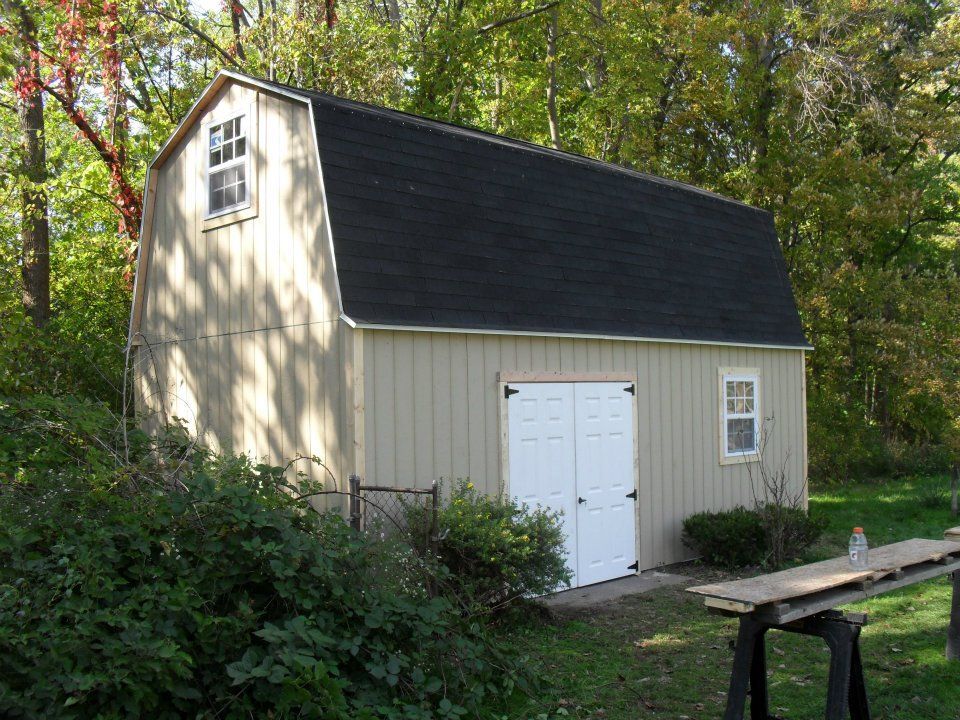 Tan barn with a black roof, white double doors, and small windows, set in a yard with trees.
