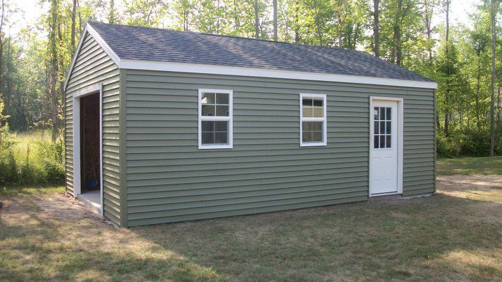 Green shed with a garage door, two windows, and a white door on a grassy lot.
