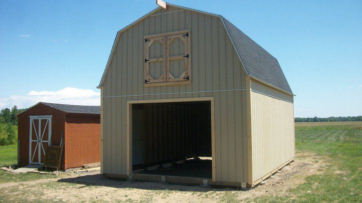 Beige barn with an open doorway, brown shed to the left, set in a field with blue sky.