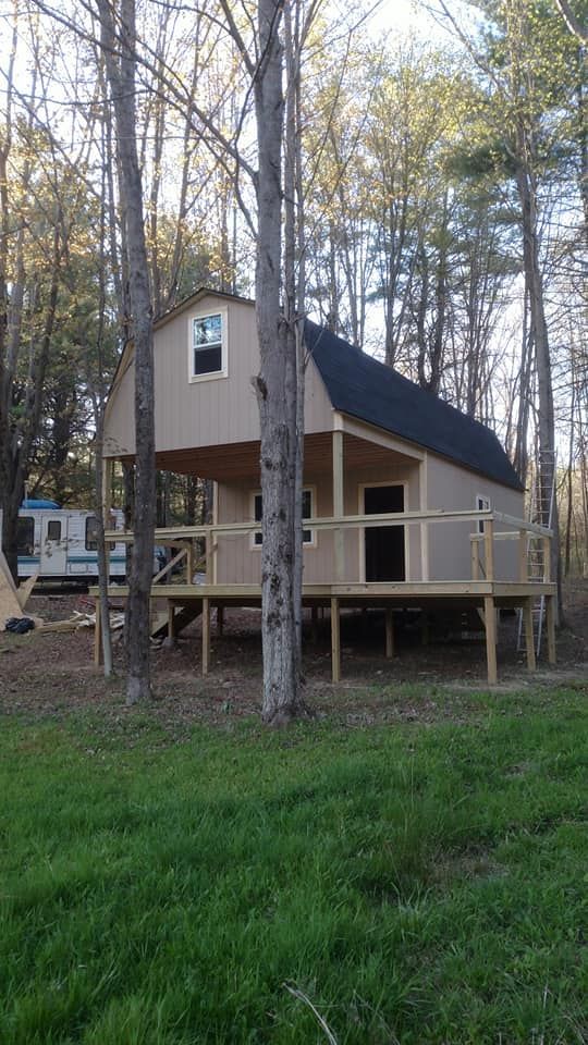 A light brown cabin with a black roof and porch, elevated on wooden supports, set in a wooded area.