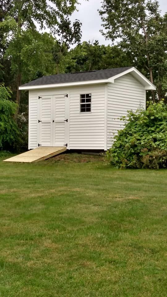 White shed with black roof, small window, and wooden ramp on green lawn.
