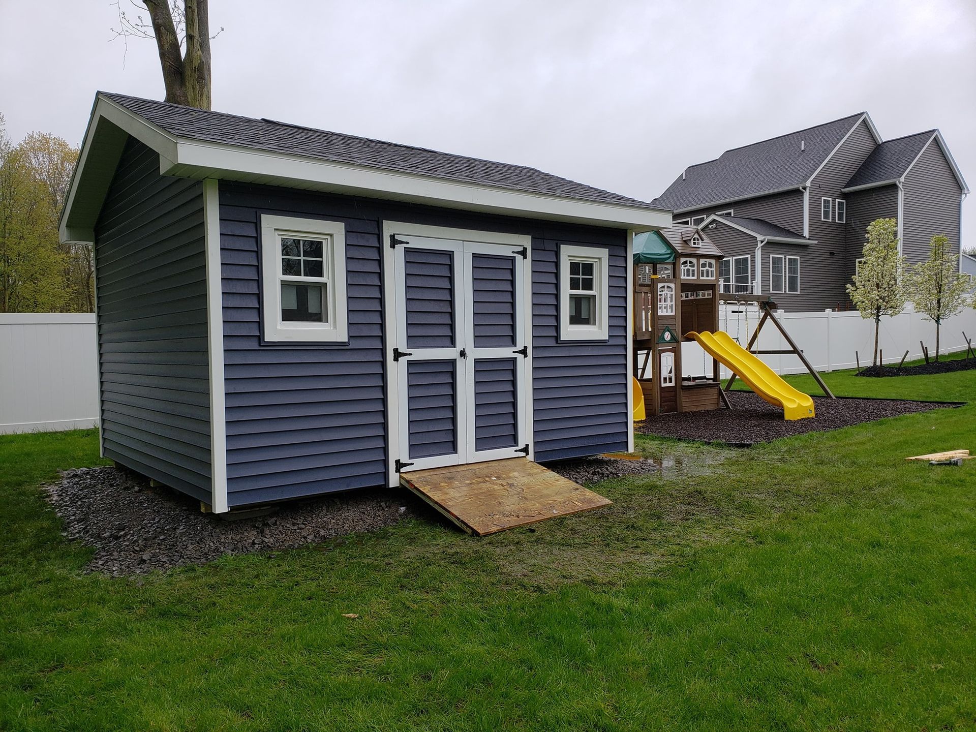 Blue shed with white trim, ramp, two windows, and double doors.  Play set and white fence in background.