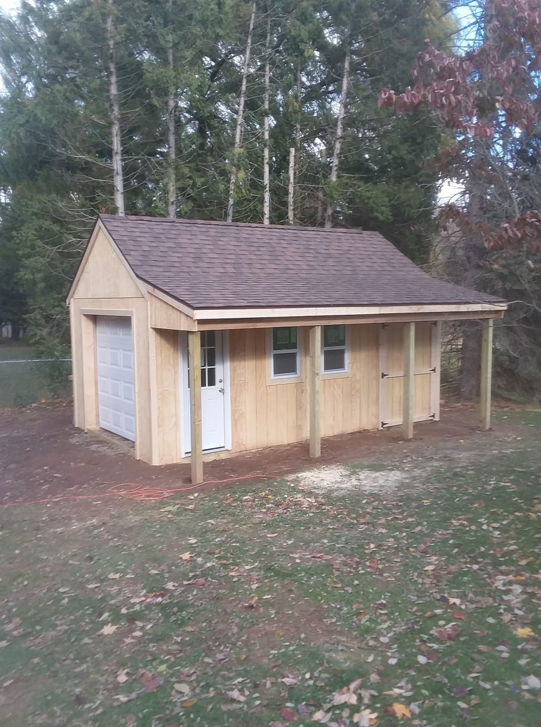 Wooden shed with garage door, white door, windows, and covered porch on a grassy lot.