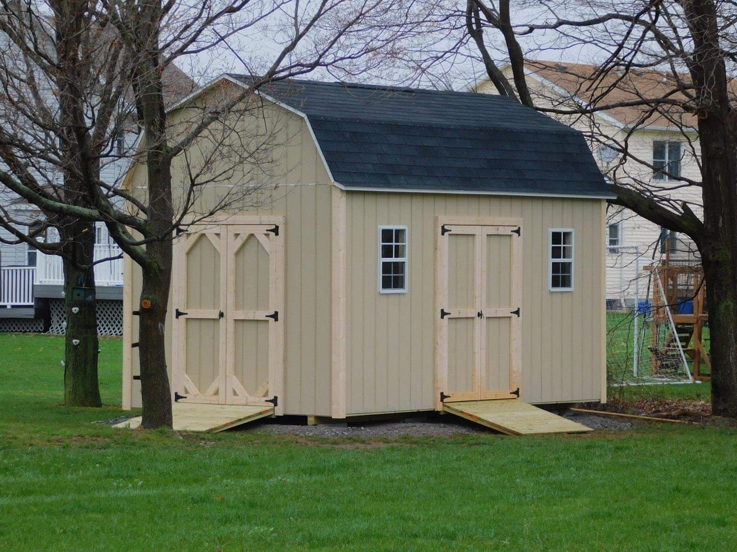 Tan shed with black roof, two doors, two small windows, wooden ramps, in a grassy yard.