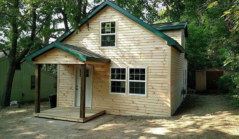 Small, light-wood house with a green roof and porch. Front door, windows, and surrounding trees are visible.